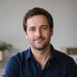 Portrait de Ambroise Marchessault, cheveux bruns courts, barbe naissante, chemise bleu nuit, sourire discret, fond clair flou