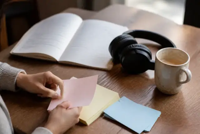 Table de café en bois avec carnet de vocabulaire ouvert, fiches bristol de couleurs douces, casque audio et tasse, une main retourne une fiche
