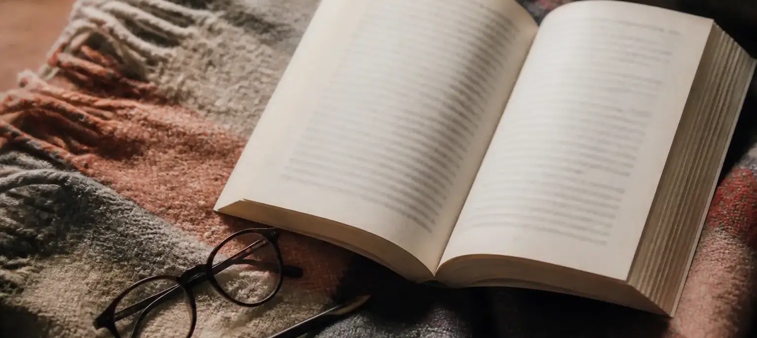 Un livre de poche ouvert posé sur un plaid, une paire de lunettes et un crayon à côté