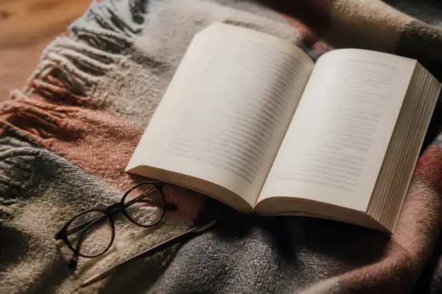 Un livre de poche ouvert posé sur un plaid, une paire de lunettes et un crayon à côté
