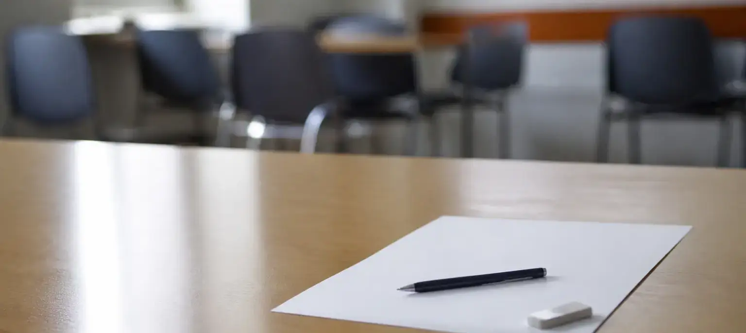 Une feuille blanche, un stylo et une gomme sur une table claire, des chaises floues au fond de la salle