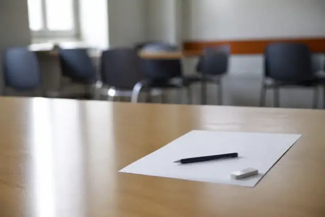 Une feuille blanche, un stylo et une gomme sur une table claire, des chaises floues au fond de la salle