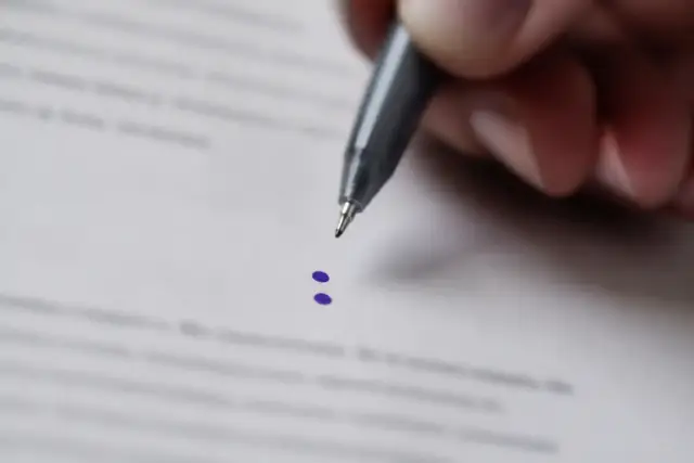 Deux points superposés tracés à l’encre violette sur une feuille blanche, entre deux lignes de texte