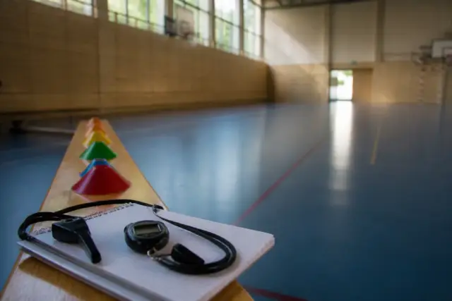 Un sifflet, un chronomètre et des plots de couleur posés au bord d’un gymnase vide, dans la lumière du matin