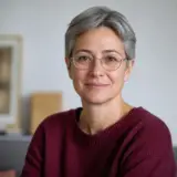 Portrait de Hortense Valleray, cheveux gris courts, lunettes fines, pull bordeaux, léger sourire, fond clair flou