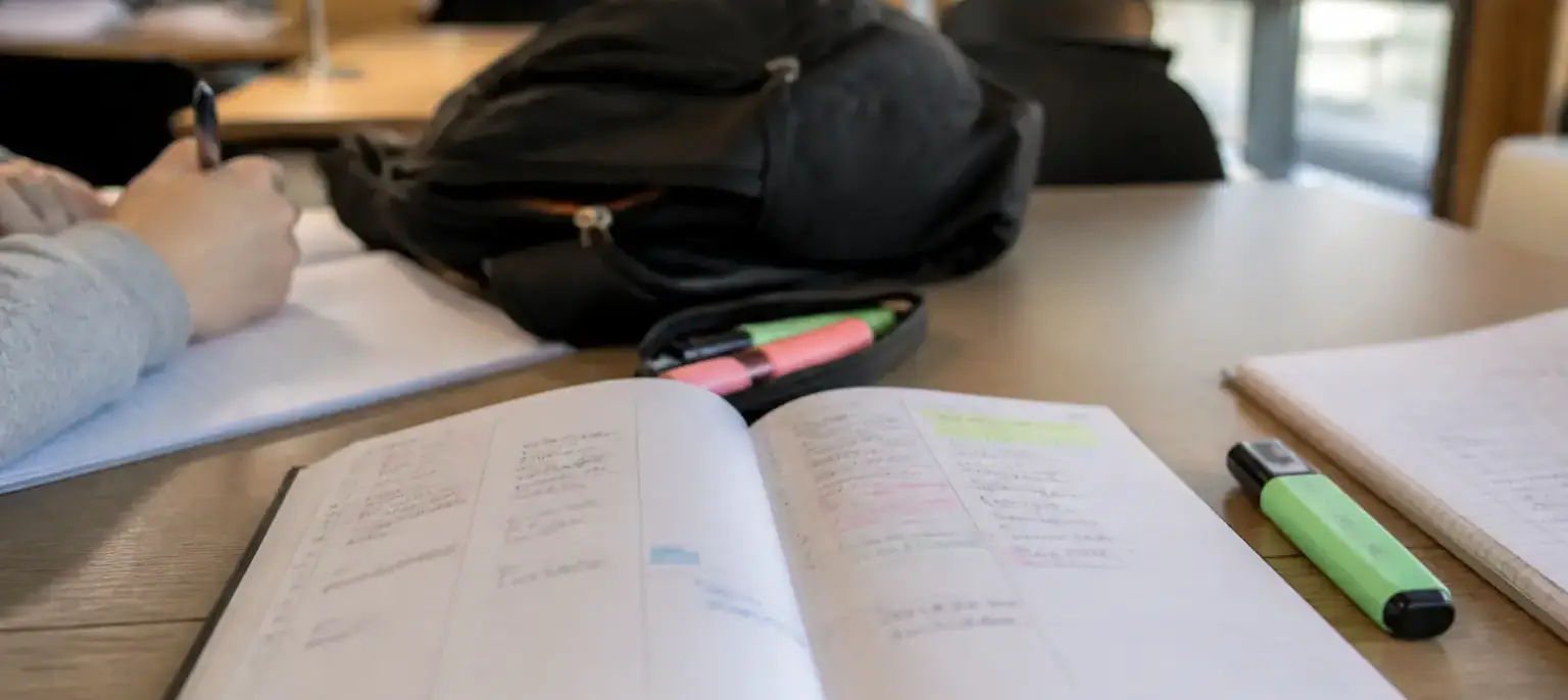 Un agenda papier ouvert sur une semaine, un surligneur et un sac à dos posés sur une table de bibliothèque universitaire
