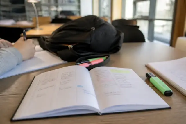 Un agenda papier ouvert sur une semaine, un surligneur et un sac à dos posés sur une table de bibliothèque universitaire